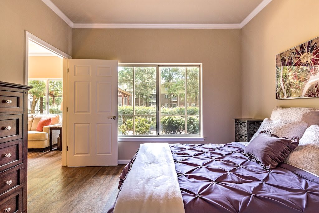 A cozy bedroom with a large window overlooking a garden, featuring a bed with purple bedding and an open door leading to another room.
