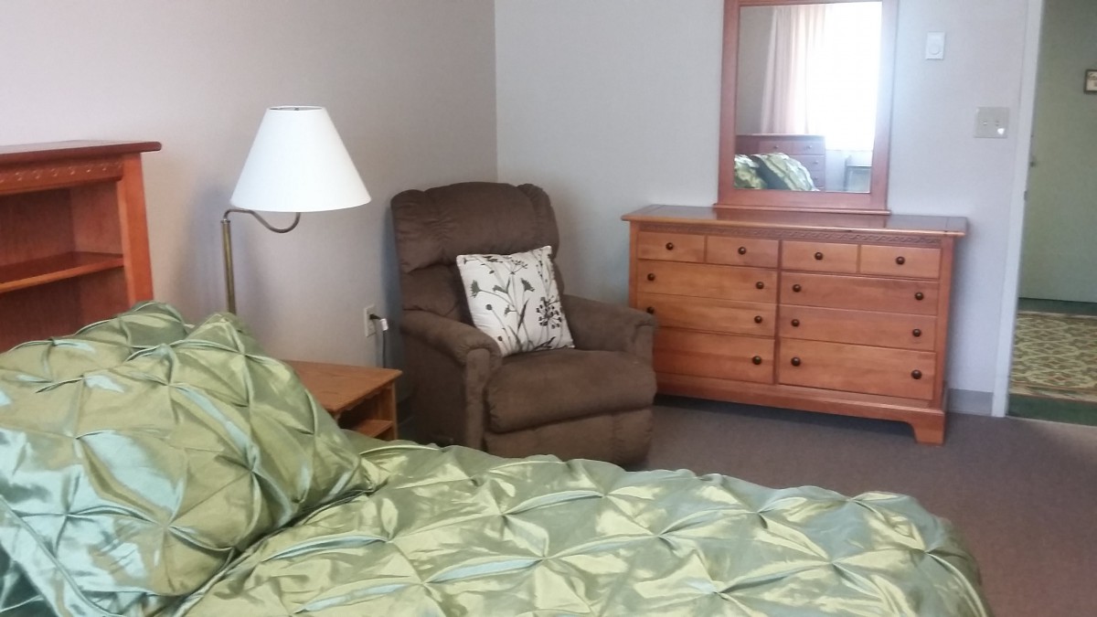 A cozy bedroom featuring a green bedspread, an armchair with a decorative pillow, a wooden dresser with a mirror, and a lamp.
