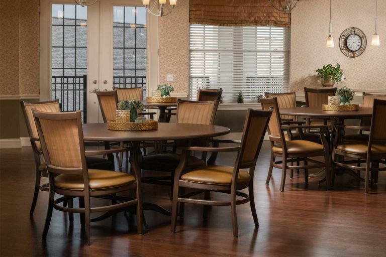 A well-lit dining area featuring a round table surrounded by chairs, with large windows and hardwood flooring.