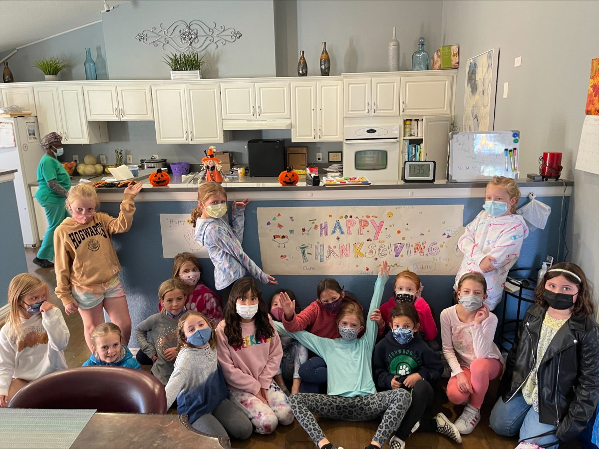 A group of children wearing masks gather indoors for a celebration in front of a decorated banner in a kitchen setting.