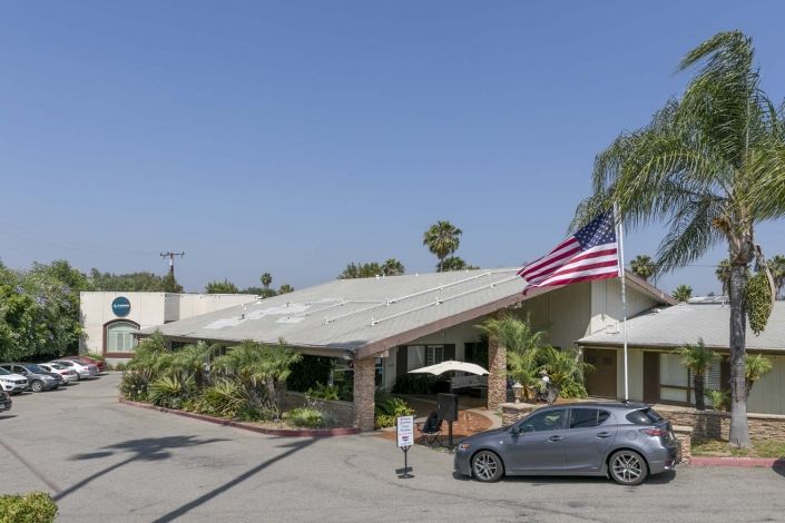 A building with a parking lot, car, and American flag surrounded by palm trees under a clear sky.