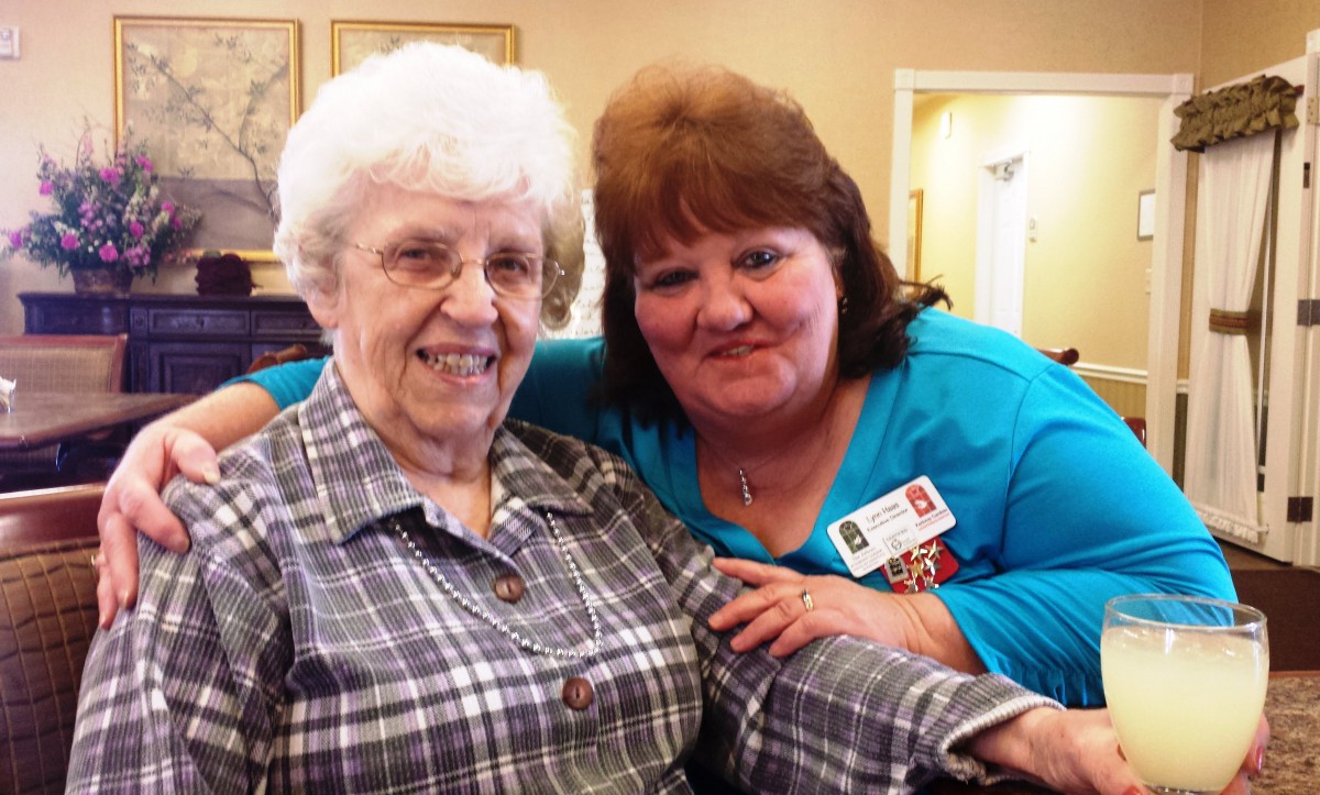 An elderly woman and a caregiver smiling together indoors.