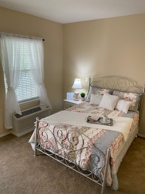 A cozy bedroom with a neatly made bed, featuring a quilt and pillows, a nightstand with a lamp, and natural light coming through the window.