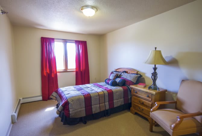 A cozy bedroom with a bed, side table, and lamp next to a window with red curtains.