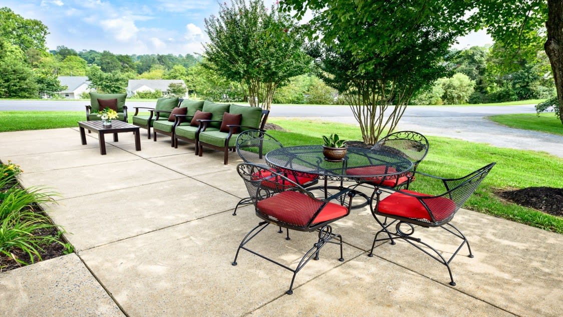 A charming outdoor patio setup featuring a round table with red-cushioned chairs and a line of sofas with green cushions, set against a lush backdrop of trees and grass.