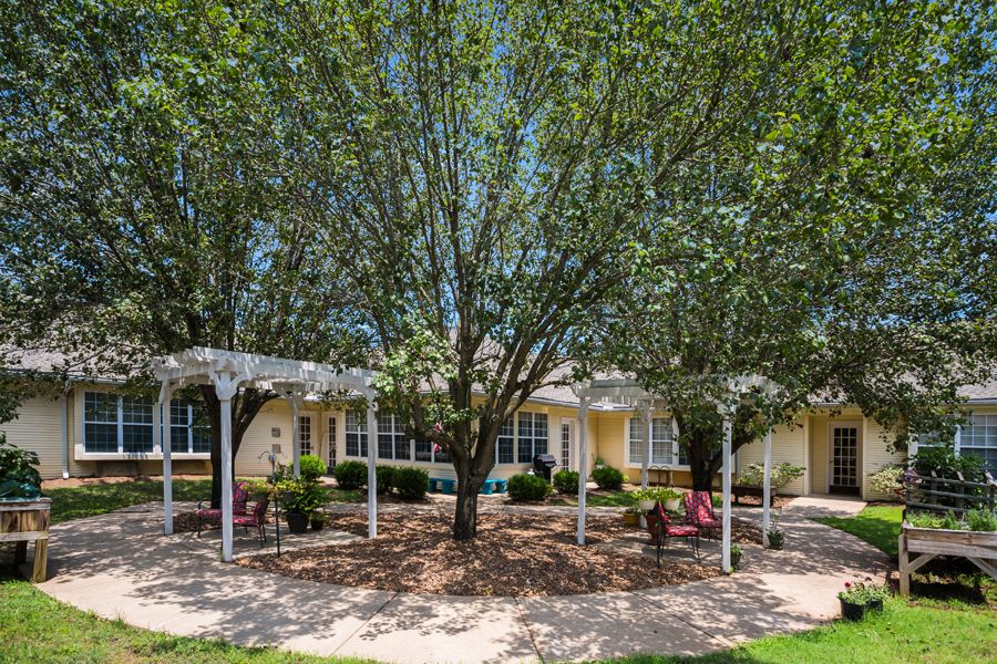 A scenic outdoor area featuring a garden with trees, chairs, and a pergola in front of a building.
