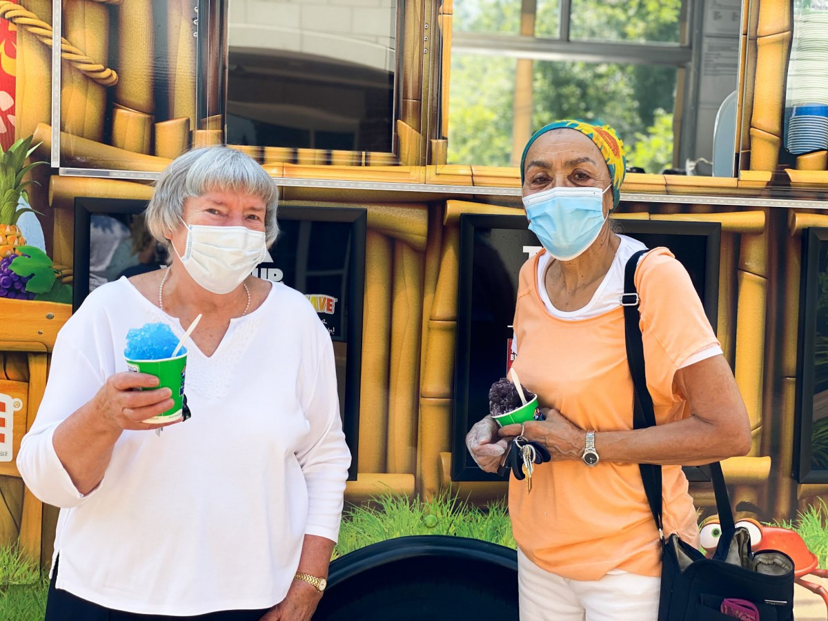 Two people wearing masks enjoy shaved ice outside on a sunny day.