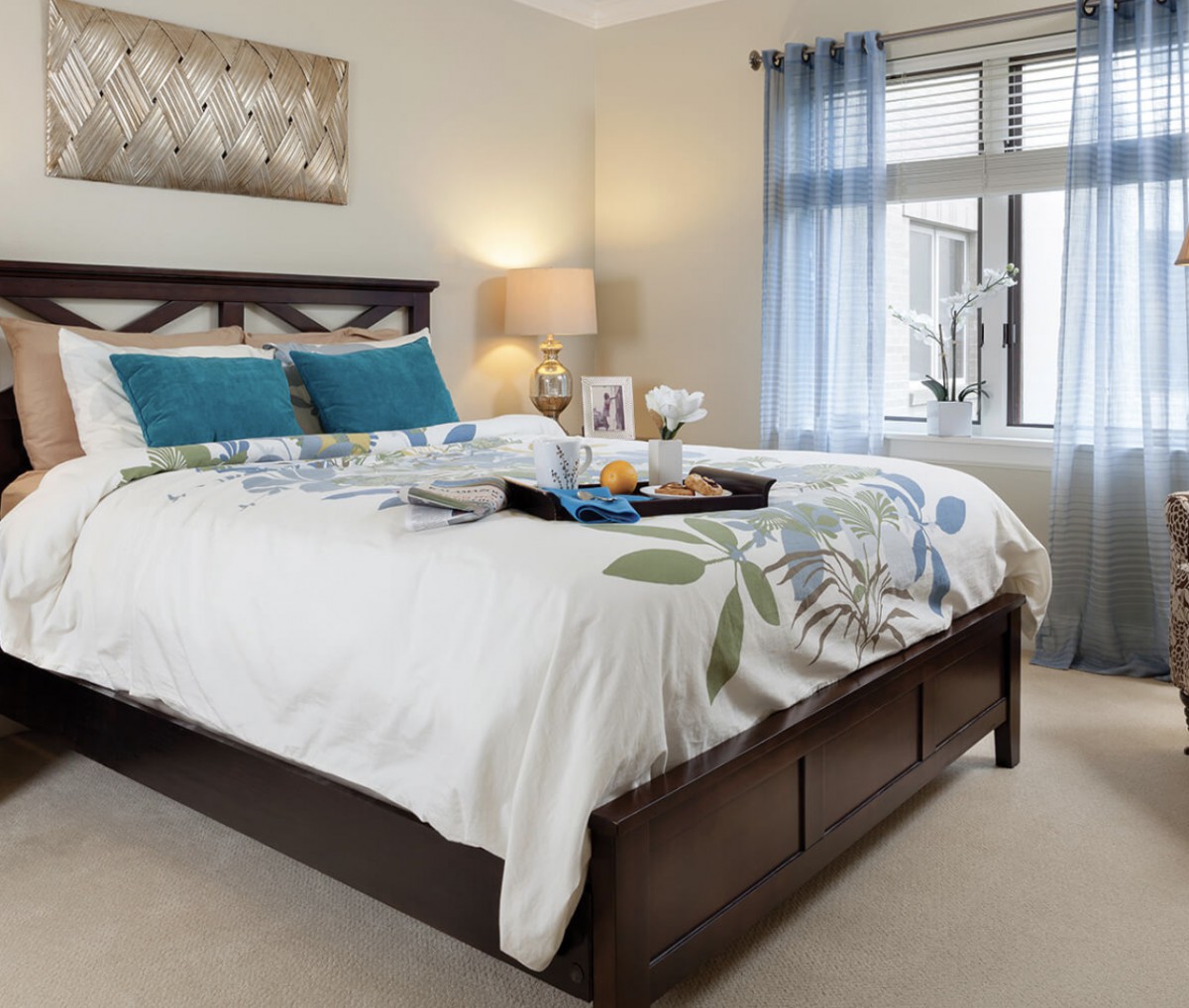 A cozy and well-decorated bedroom featuring a large bed with pillows, a bedside lamp, and a window with blue curtains.