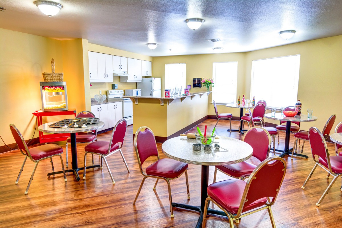 A bright and inviting dining area with red chairs and white tables, adjacent to a kitchen space.
