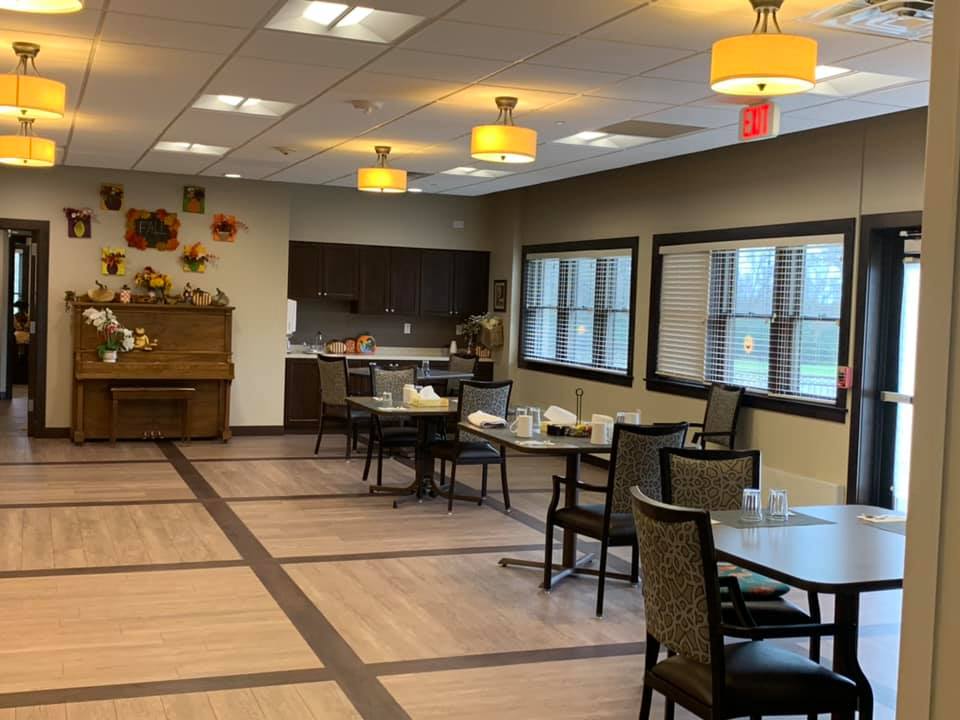 An empty dining room with neatly arranged tables and chairs, featuring a kitchen area and large windows.