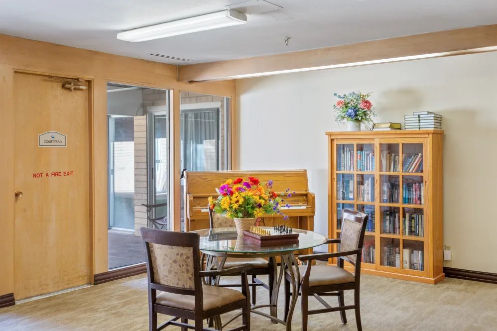A cozy room with a round table and chairs, surrounded by a piano and a bookshelf filled with books.