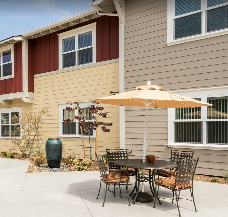 A cozy outdoor patio area with a table, chairs, and umbrella in front of a multi-colored residential building.