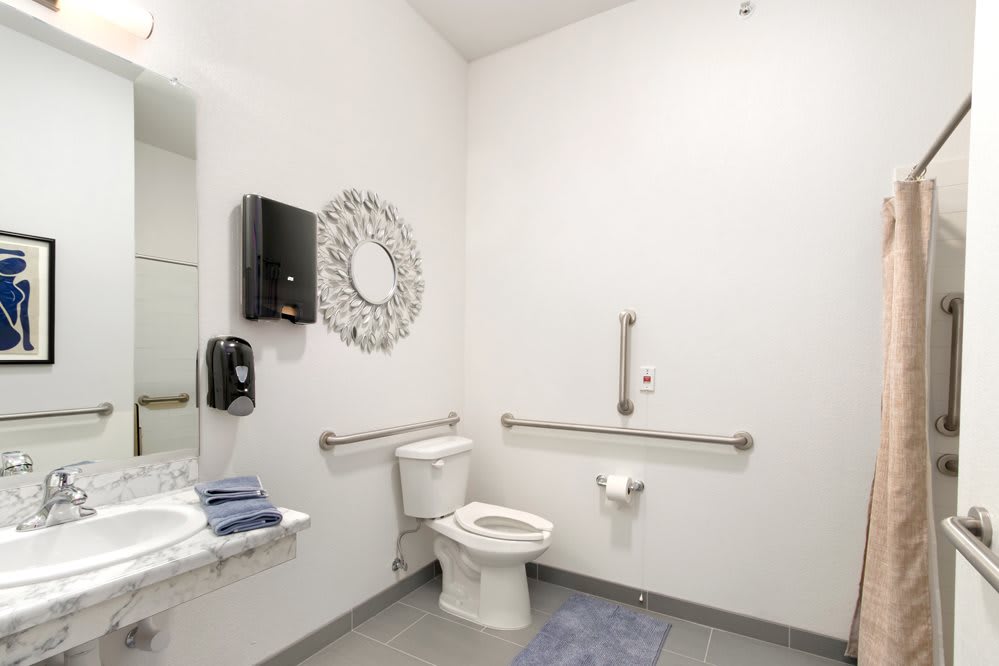 An accessible bathroom with a toilet, handrails, and a wall-mounted mirror next to a sink.