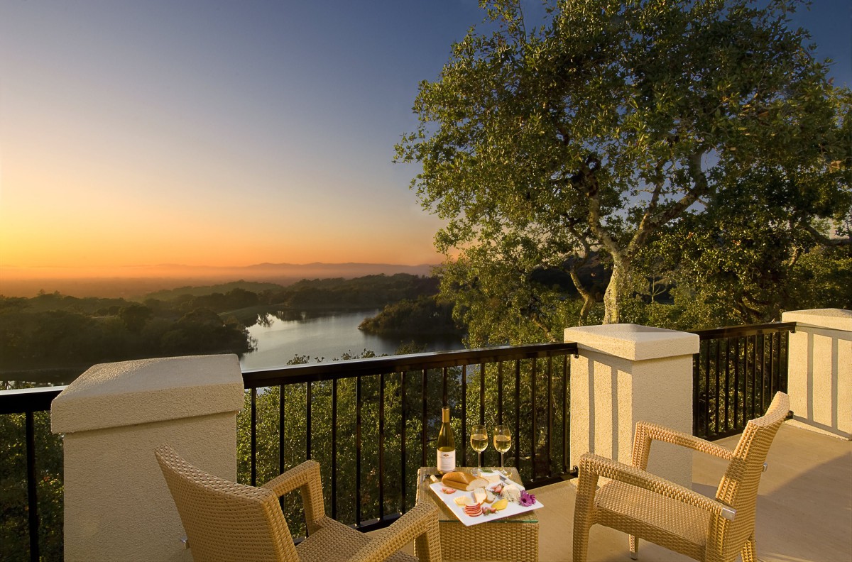 A serene sunset view from a balcony, featuring a table set with wine and chairs overlooking the landscape.
