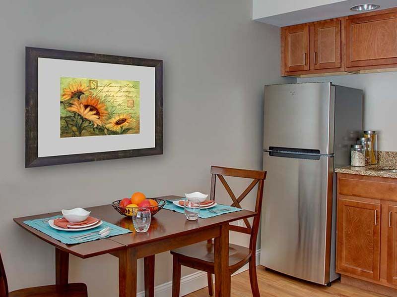 A cozy kitchen nook with a dining table set for two, featuring a sunflower painting and a modern fridge.