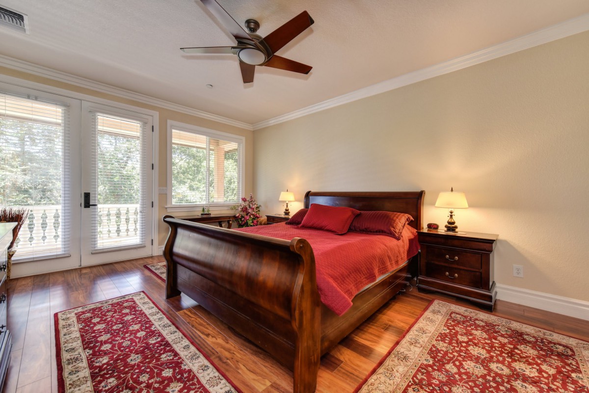 A cozy bedroom featuring a wooden sleigh bed with red bedding, complemented by matching area rugs and warm lighting from bedside lamps. Large windows allow natural light to fill the room.