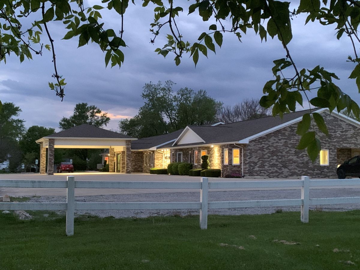 A suburban house with lights on during a cloudy evening, framed by trees and a white fence.