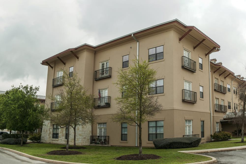A modern three-story apartment building with balconies, surrounded by trees and grass.