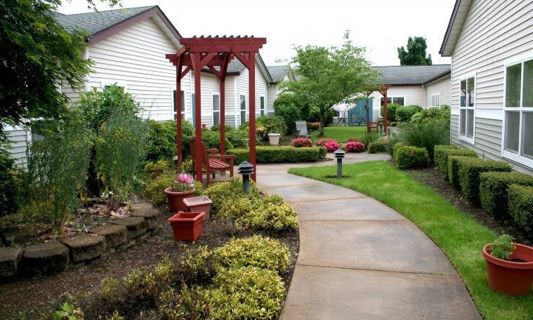 A beautifully landscaped garden with a winding pathway, red arbor, and benches surrounded by lush greenery and shrubs.