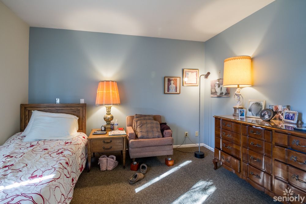 A cozy bedroom with a bed, nightstand, and comfortable chair illuminated by warm lights.