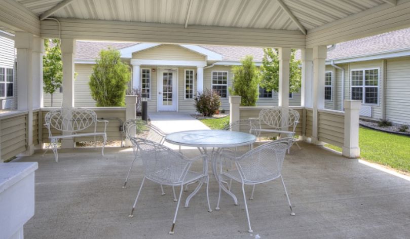 A sunny patio with outdoor furniture under a gazebo, overlooking a garden and house.
