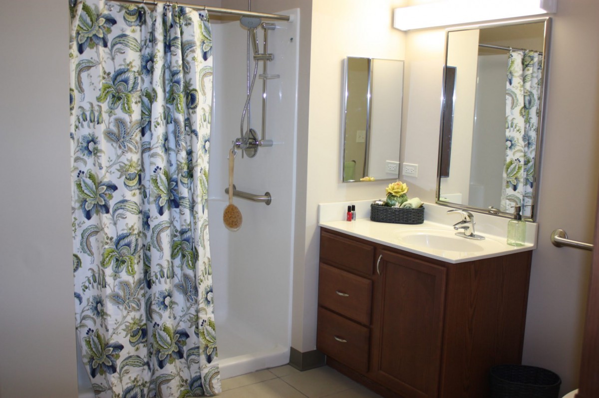 A well-lit bathroom featuring a shower with a floral curtain, a sink with cabinets, and a mirror.