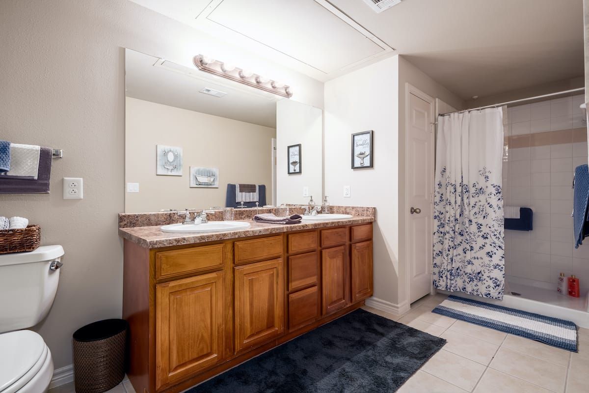 A well-lit bathroom featuring a double sink vanity with wooden cabinets, a large mirror, and a patterned shower curtain.