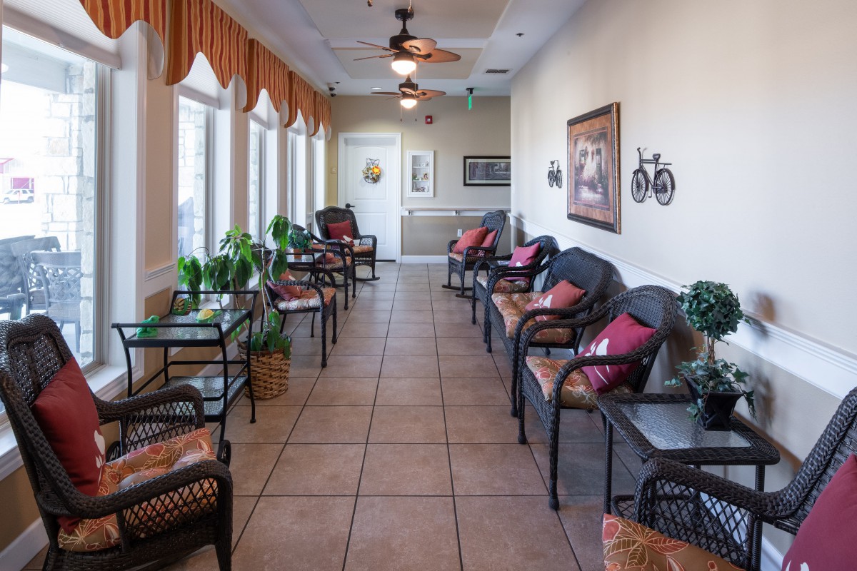 A cozy waiting room with multiple chairs and tables lined up against a bright, windowed wall, adorned with plants and decorative items.