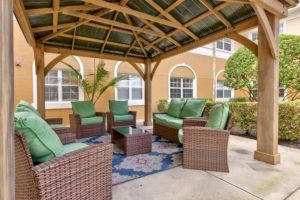 A cozy outdoor seating area under a gazebo, featuring wicker chairs with green cushions and a coffee table on a colorful rug.