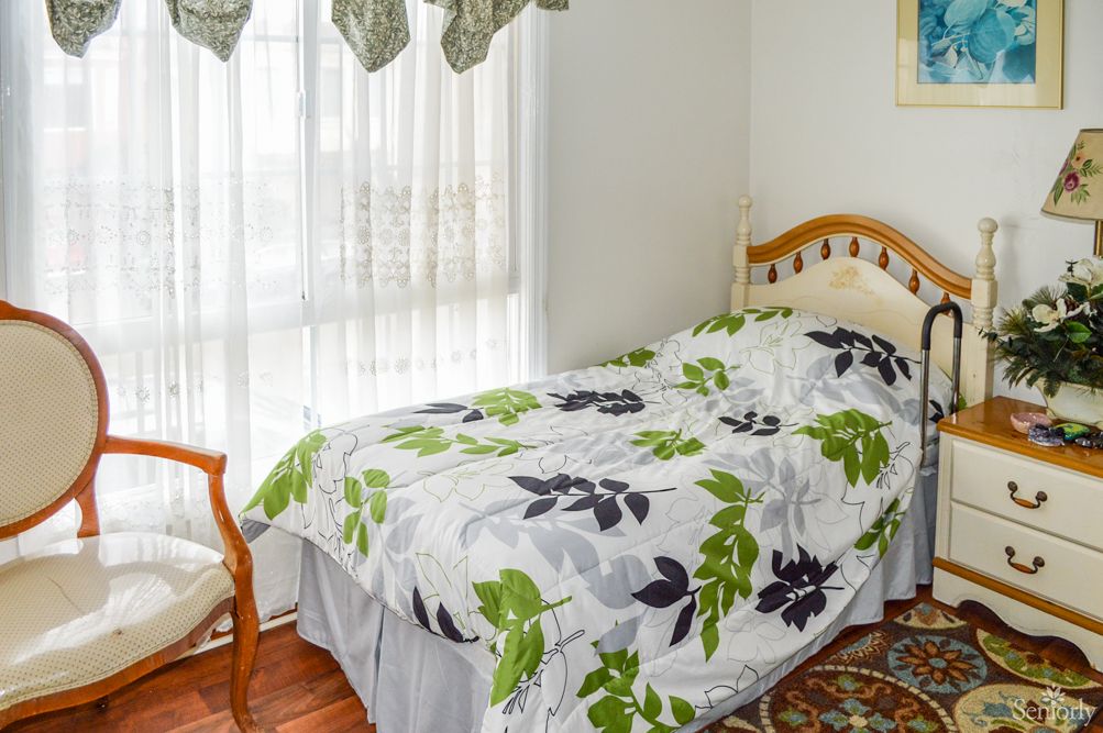 A cozy bedroom featuring a single bed with floral bedding, a chair, and large window with curtains.