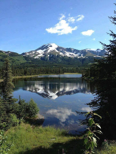 A serene view of a mountain reflected in a tranquil lake, surrounded by lush greenery and a clear blue sky.