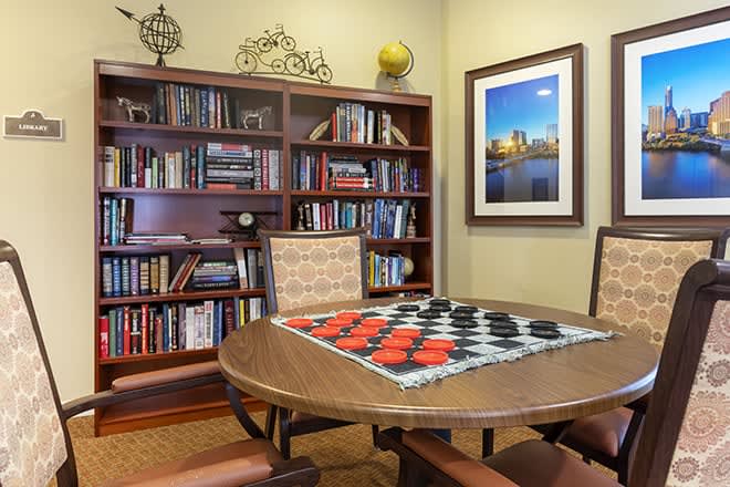 A cozy library room with bookshelves, a table set for a game of checkers, and framed artwork on the wall.