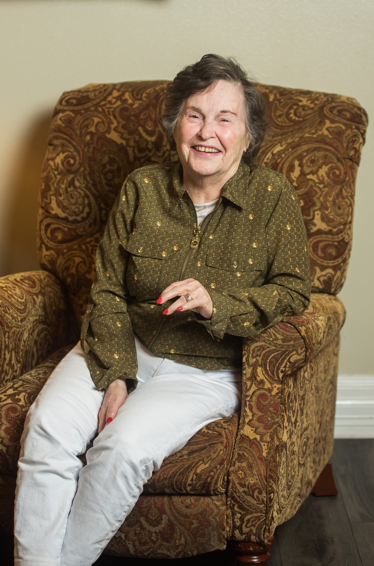 An elderly woman smiling while sitting on a comfortable chair indoors.
