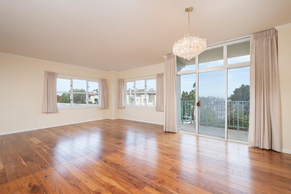 A spacious, empty room with hardwood flooring, large windows, and a chandelier, offering plenty of natural light and access to a balcony.