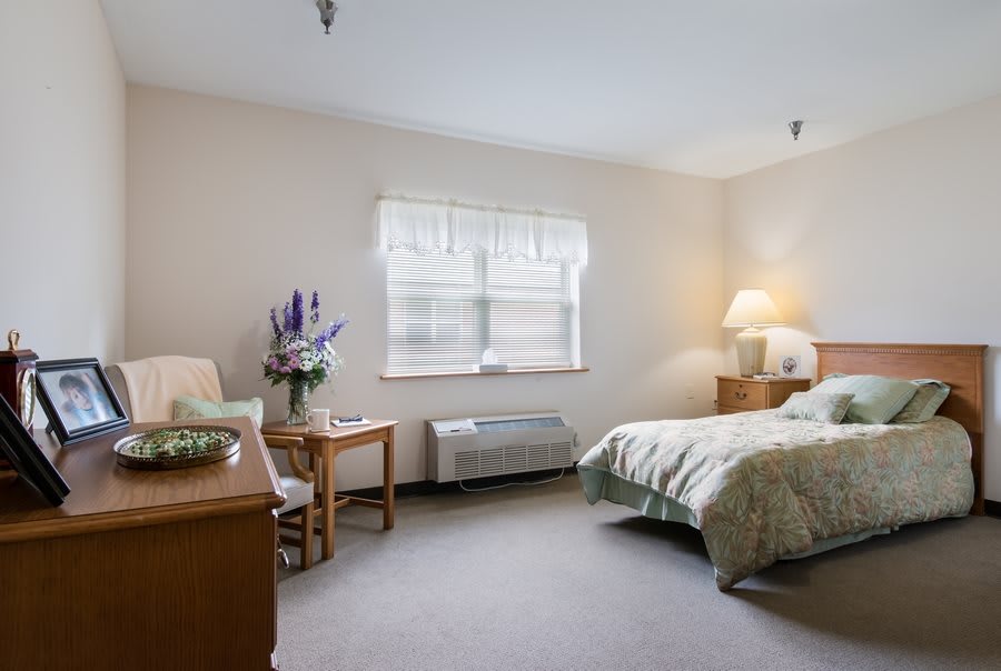 A cozy bedroom with a neatly made bed, a table and chair, and a warm lamp on the nightstand.