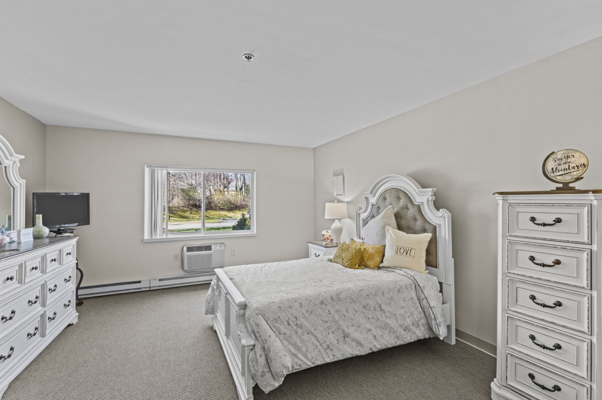 A spacious bedroom with a neatly made bed, neutral tones, and natural light coming through a large window.