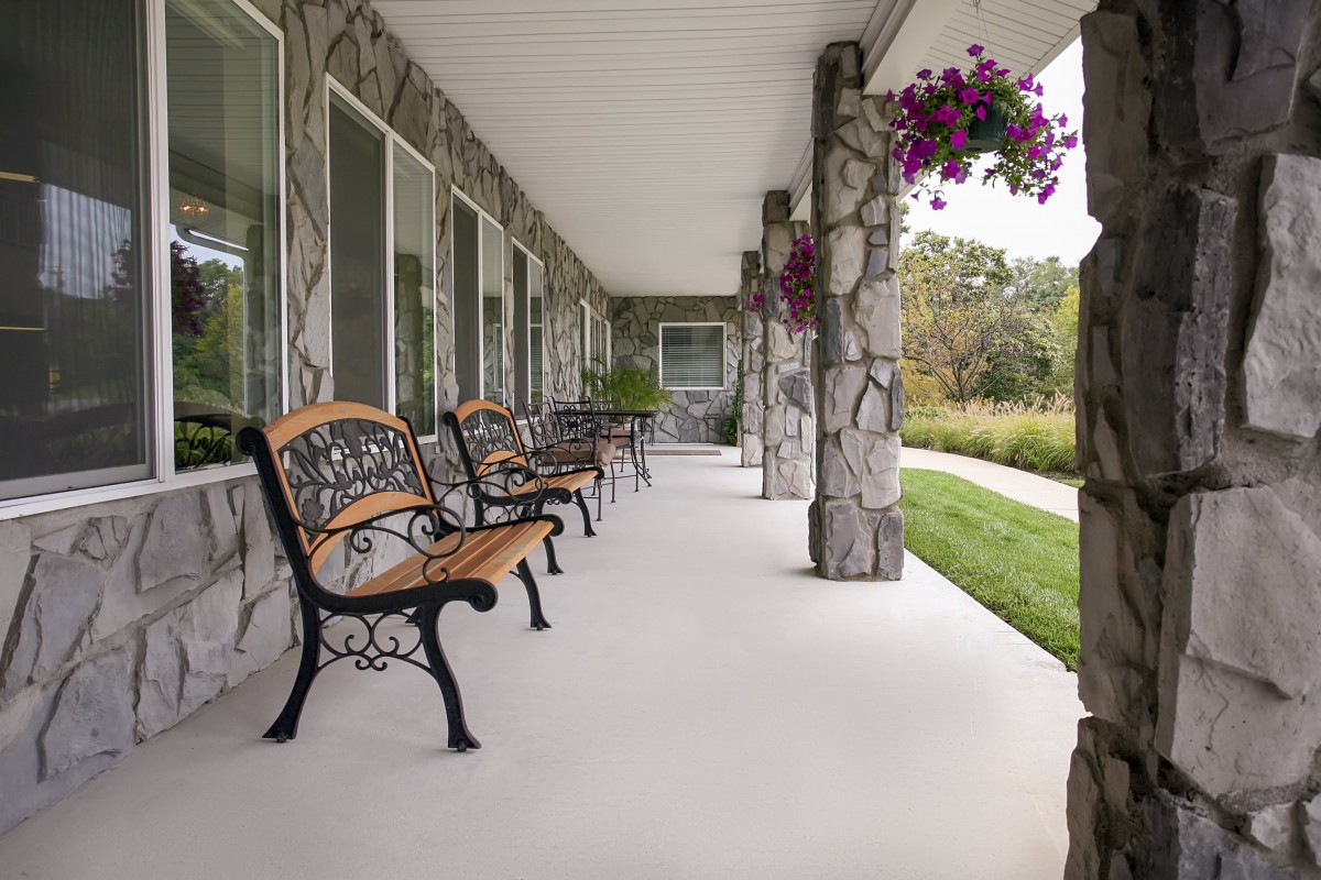 A porch with stone columns and benches, adorned with hanging flowers and surrounded by greenery.