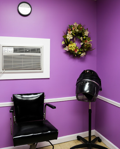 A cozy hair salon corner featuring a black chair, a vintage hair dryer stand, and vibrant purple walls adorned with a floral wreath.