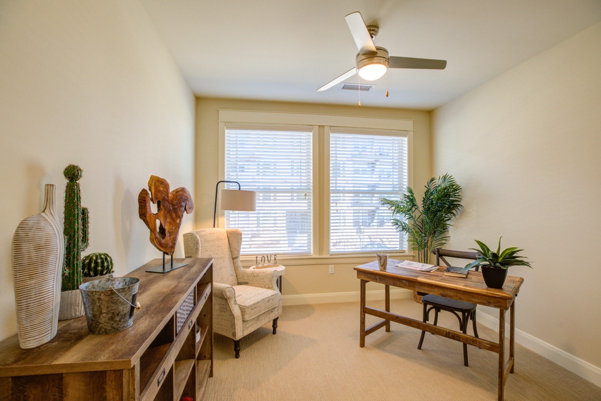 A cozy home office with a wooden desk, chair, bookshelf, and large windows providing natural light.