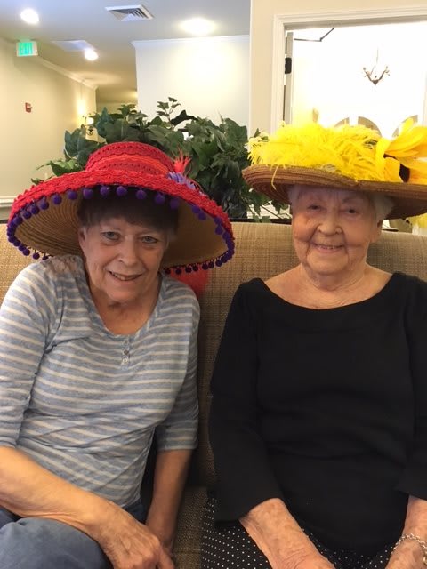 Two elderly women smiling while wearing colorful hats indoors.