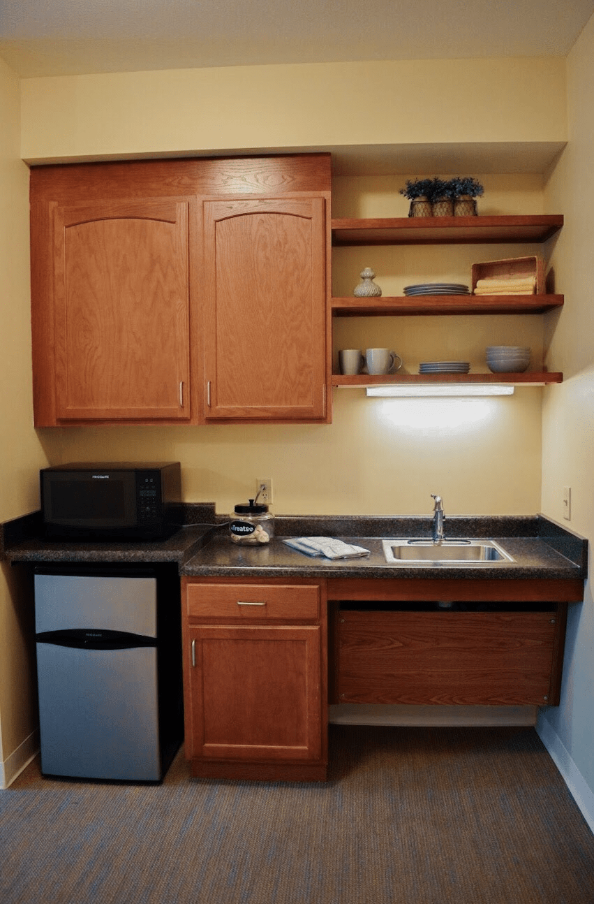 A small kitchen area with wooden cabinets, open shelves, a microwave, mini fridge, and sink.