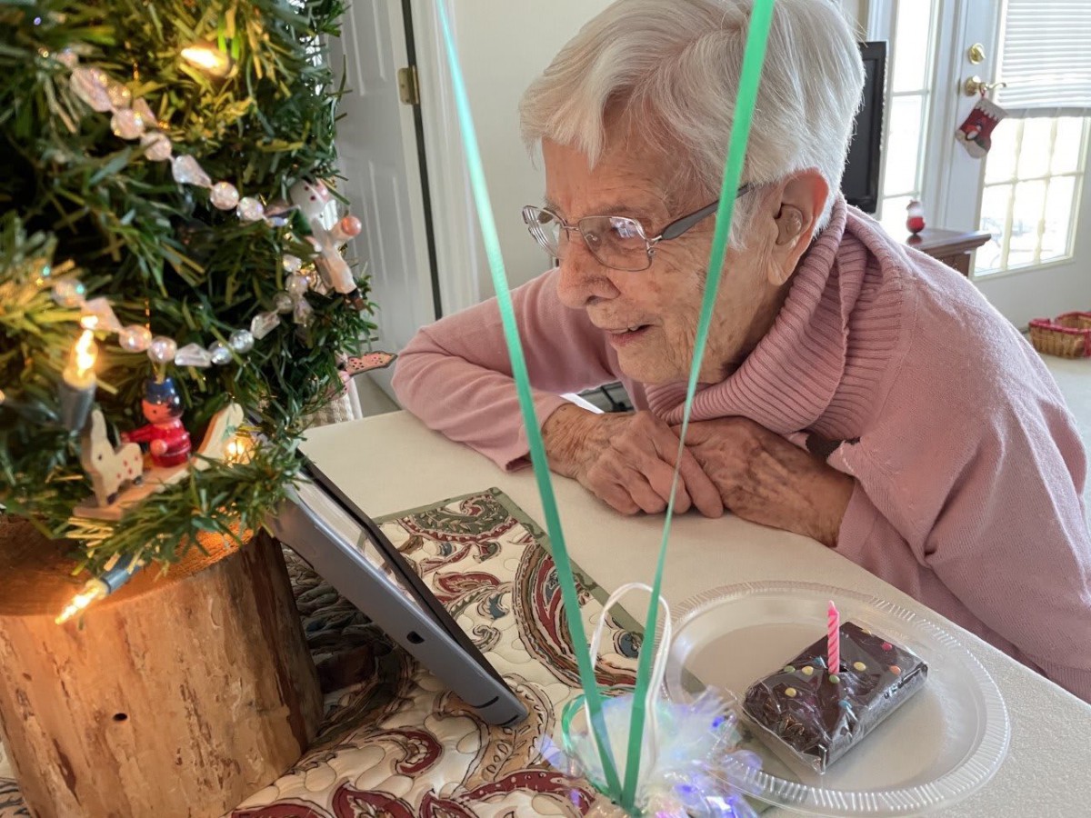 An elderly woman smiling during a video call, celebrating with a small birthday cake and Christmas decorations.