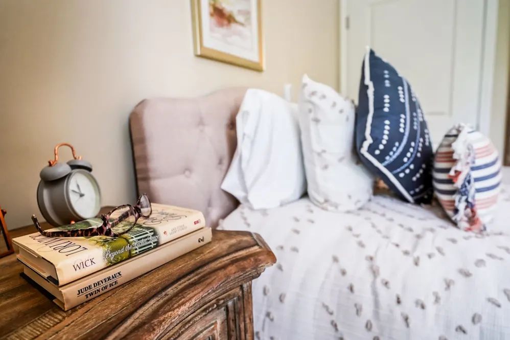 A cozy bedroom with a neatly made bed, featuring decorative pillows and a rustic nightstand topped with books and a lamp.
