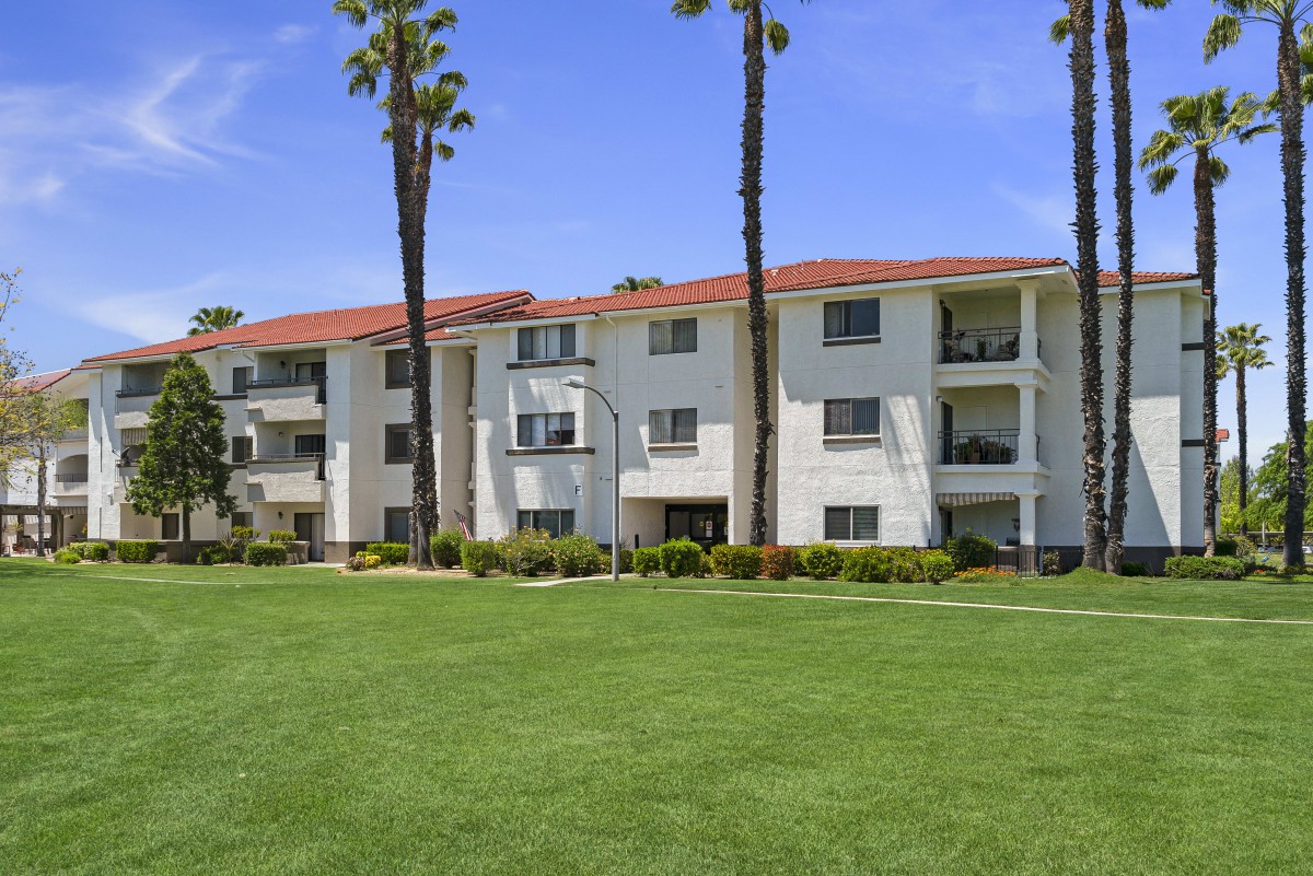 A three-story apartment building surrounded by a well-maintained lawn and tall palm trees on a sunny day.