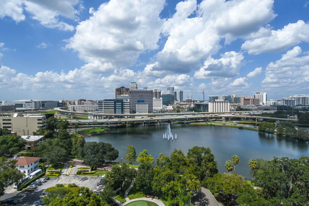 A vibrant cityscape with a lake and park in the foreground under a sky filled with clouds.