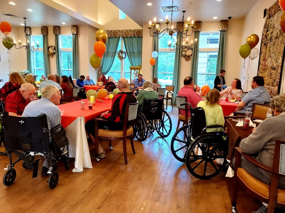 A group of seniors enjoy a social gathering in a decorated dining room, with some attendees seated in wheelchairs.