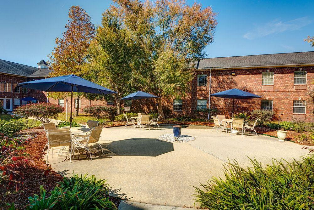 A sunny patio area with tables, chairs, and large umbrellas surrounded by trees and a building.