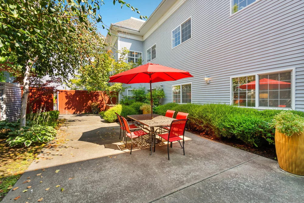 A sunny patio area with a red umbrella and matching outdoor furniture, surrounded by greenery.