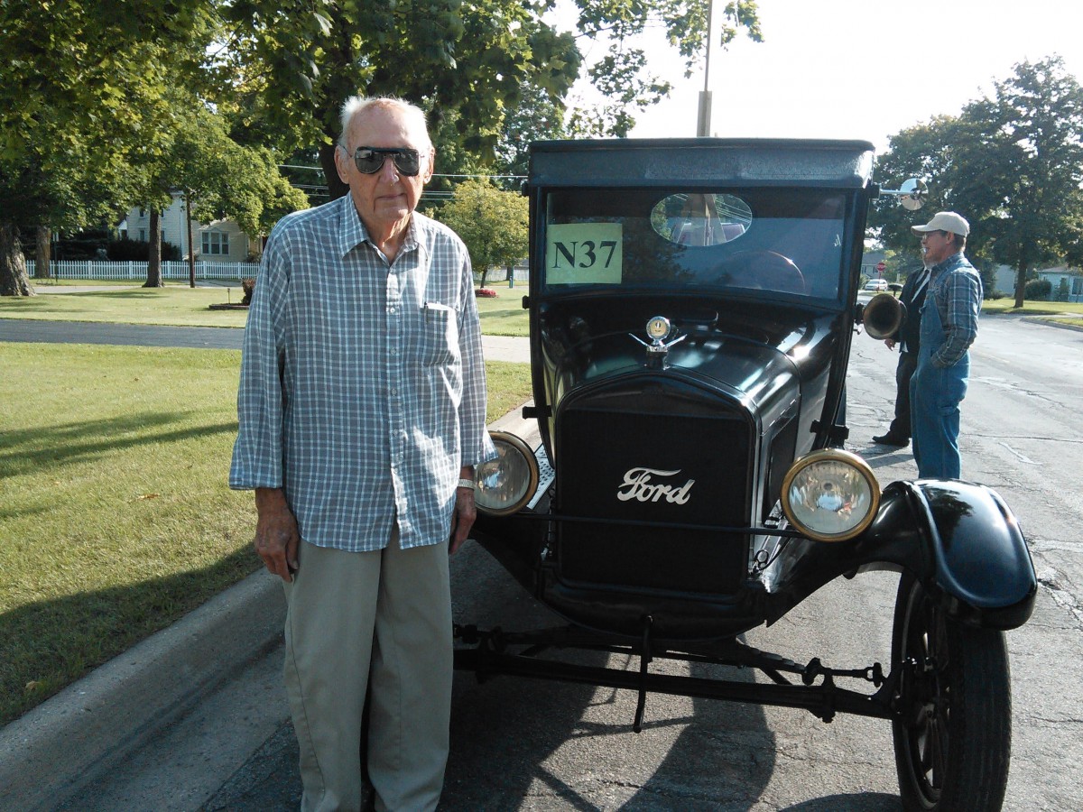 An elderly man stands beside a vintage Ford car on a sunny day.