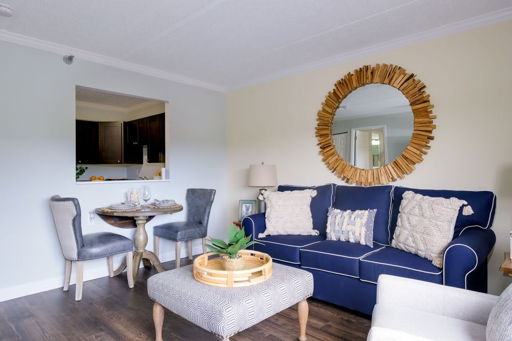 A cozy living room with a blue sofa, decorative pillows, and a round mirror above. A small dining table with chairs is visible in the background.